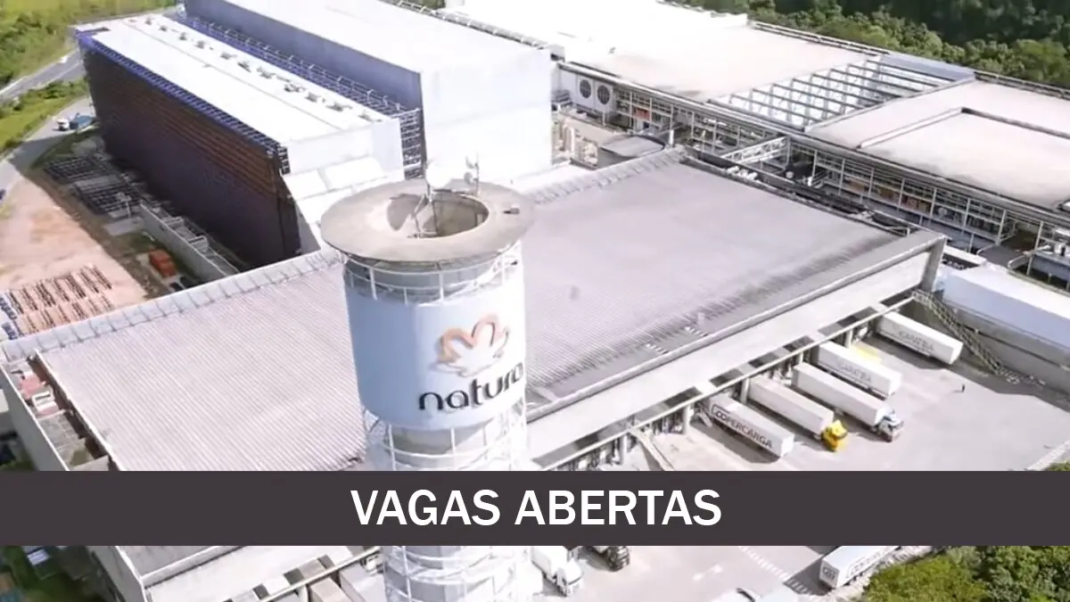 Vista aérea do prédio da Natura &Co em Cajamar, SP, com caminhões estacionados na frente. A imagem também mostra a placa "VAGAS ABERTAS" indicando oportunidades de emprego na empresa.